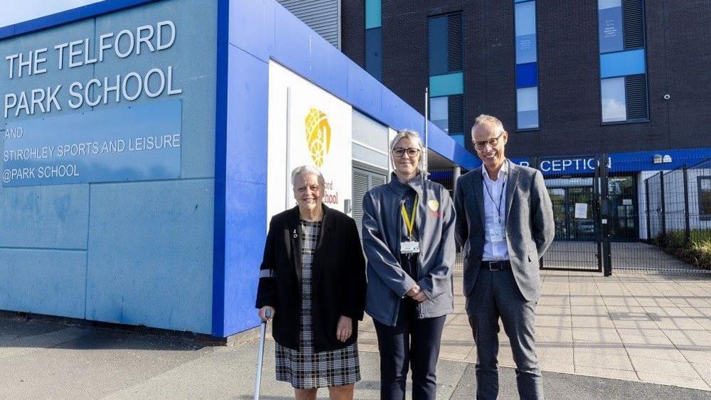 Two women and a man are standing in front of a school building. the woman on the left has white hair and a black jacket, with a stiped dress and a crutch. The woman in the middle is wearing a grey-blue jacket with a school logo on it, and a yellow lanyard. The man has a grey suit on with black glasses and a blue lanyard. A blue building extension has a sign on it reading "The telford park school"