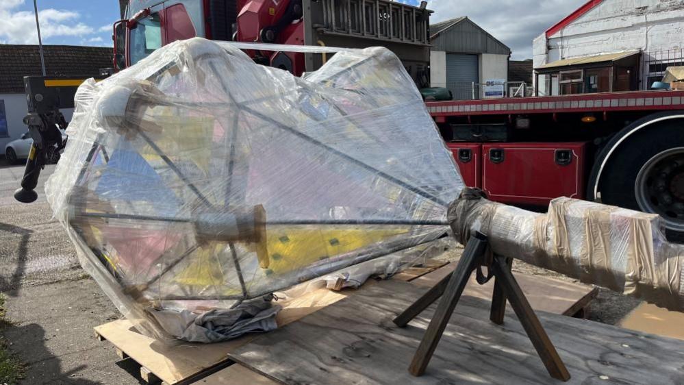 A steel column with a steel frame forming a heart shape, filled with glass and steel plates. The sculpture is wrapped in clingfilm and is on a pallet having been craned off a lorry, in the backdrop.