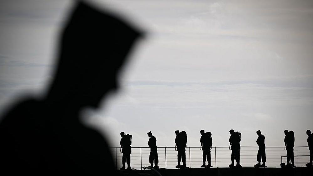 A close-up shot on a silhouetted figure of a soldier in the foreground, out of focus. In the background, in focus, eight more figures stand atop a wall by a waist-high metal fence.