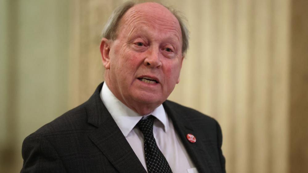 Jim Allister looks away from the camera as he speaks. He is balding and has grey hair at the sides. He is wearing a black suit, white shirt and black tie. He has a red button pinned on his jacket.