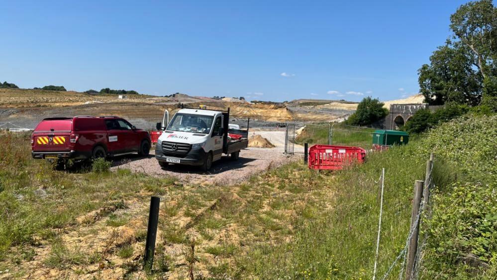 A white lorry with a door open and a red van are parked on temporary hardstanding in a grass field. There are red barriers to the right of the vehicles. There is a tree to the right and a construction site behind.
