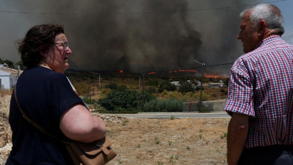A concerned looking woman with a dark brown bob, wearing a t-shirt, looks at a greying man who is wearing a checked shirt. In the distance, dark smoke is seen billowing above a line of fire going through shrubland.