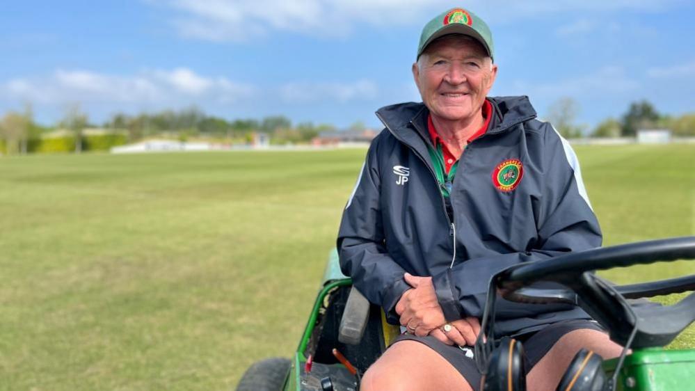 Jimmy Perchard wearing Farmers Field clothing on the top of his lawn mower in front of his cricket ground 