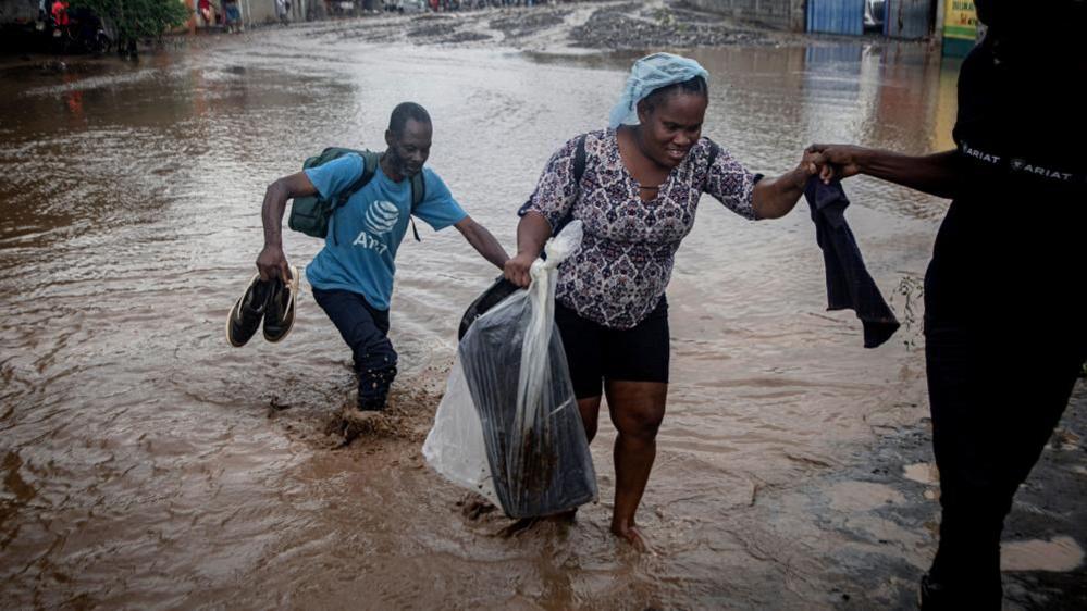 A man, wearing a blue T-shirt and carrying his shoes, follows behind a woman holding a plastic bag and being helped by another person off-frame as they wade through knee-deep muddy flood water