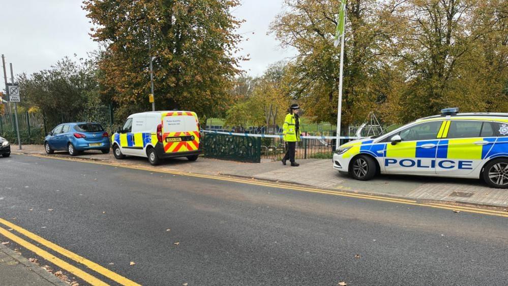 Two police cars, a police officer, by police tape, a park in the distance, with trees and foliage. Another blue car is to the left. A road is in front of the park. 