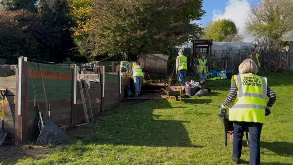 Five volunteers, dressed in jeans and luminous green, high-vis tabards, doing various jobs on site. One is pushing a wheelbarrow, another two are working by a soil tumbling machine, two others are checking the state of the compost.