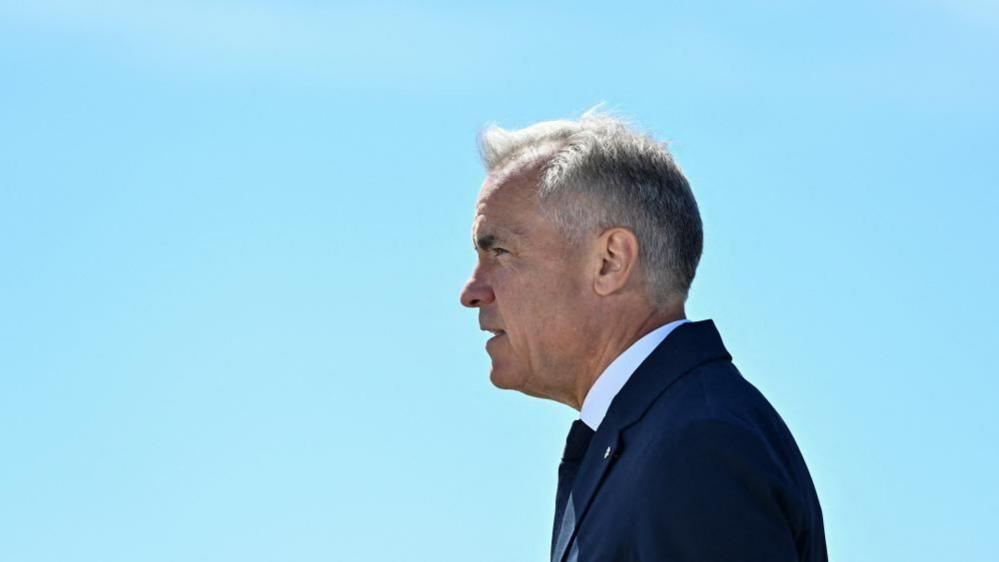 Mark Carney stands in profile, facing to the left, against a clear light blue sky. He wears a dark blue suit and tie.