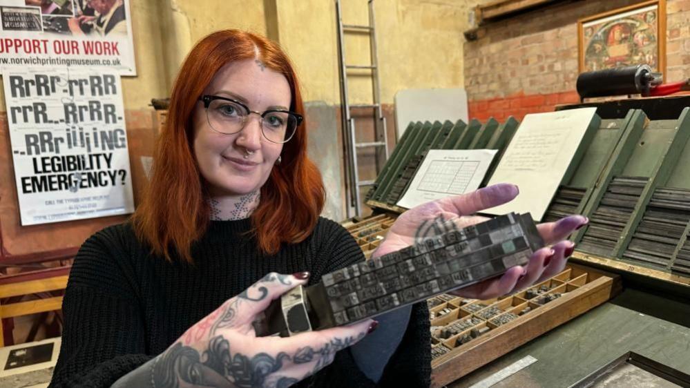 Lacey Law holds a chase of type metal near a compositor's desk. She has shoulder length ginger hair, is wearing glasses, has a nose piercing and is wearing a black round-neck jersey. Tattoos are visible on her neck, arm and both hands, with printers' ink on her left fingertips. There are racks of type metal and spacers, and a tray of type on the desk. On the back wall of the barn is a ladder and a couple of posters.
