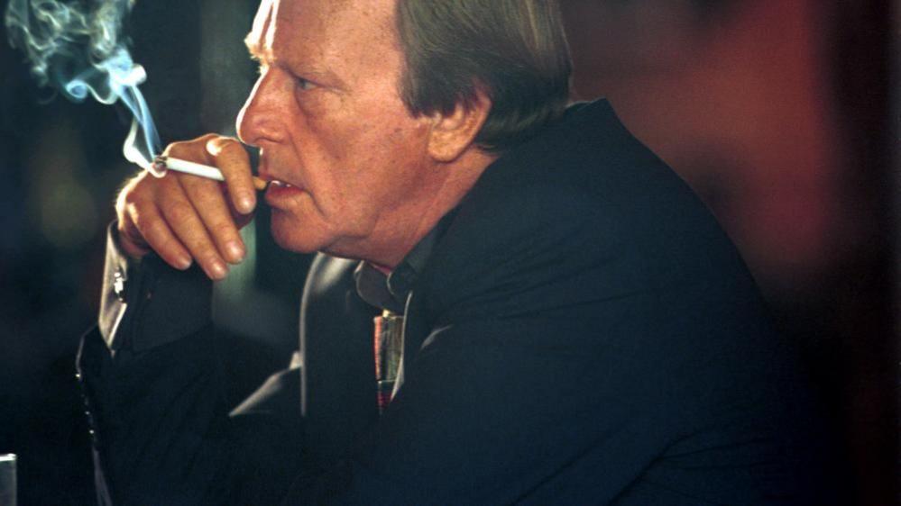 Dennis Waterman: A man with brown hair looking to his right and wearing a dark jacket. He is hold a white cigarette with a brown tip to his lips with his right hand. Smoke is issuing from the cigarette.