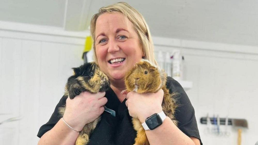 Claire McConaghy. She has blonde hair and blue eyes and wearing dark coloured scrubs, an Apple watch one her left wrist and silver bracelet on her right. Claire is holding up two guinea pigs, the one on the left is black and tan and the one on the right is mainly tan with bits on white.