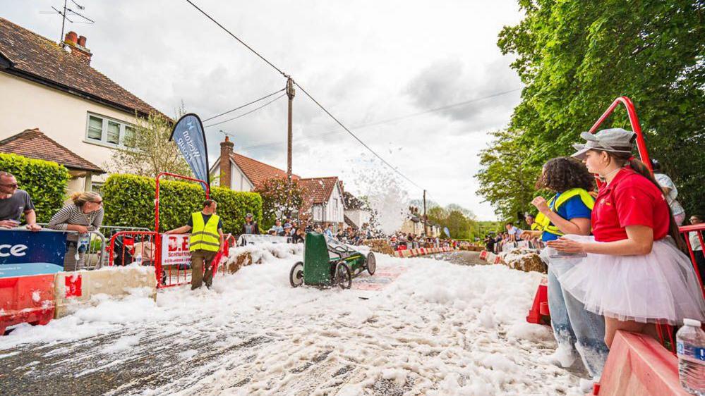 White foam is strewn across the road as a green kart races through a residential street, during last year's race. Spectators are stood at the side of the road. Some stewards are also present wearing fluorescent tabards. There are trees and bushes on either side of the road.