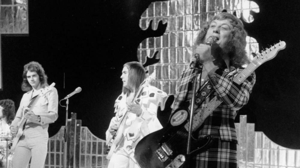 The band Slade perform on stage in the 1970s. The image is in black and white. 