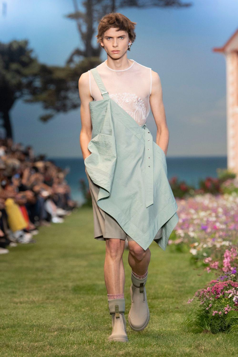A man walks down a grass runway bordered by flowers wearing a high-fashion outfit inspired by gardeners