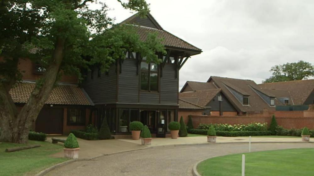 A red brick and timber-clad array of buildings, with red concrete tiled roofs. There is a driveway circling around the front entrance, with lawns and paths, and planted beds by the walls in the distance.