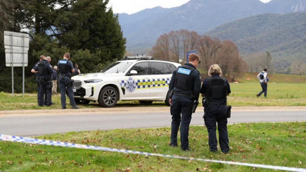 Police talking on the side of a country road, with a Highway Patrol vehicle in the background and police tape in the foreground. The landscape is green and mountainous.