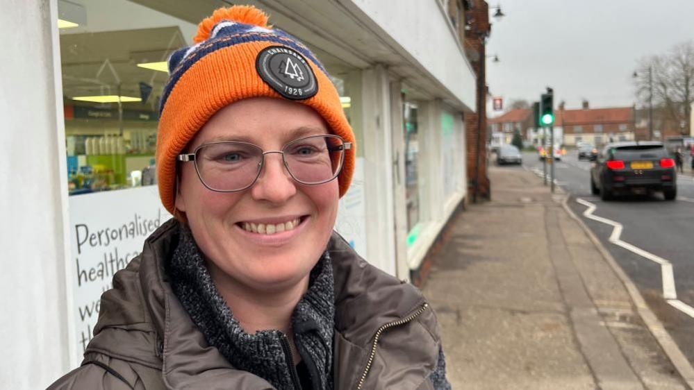 Jessica Murison is wearing a brown jacket and grey mottled jersey, and an orange and blue hat, featuring a black logo badge. She is standing outside the Acle Pharmacy on the high street. She is wearing glasses and is smiling.