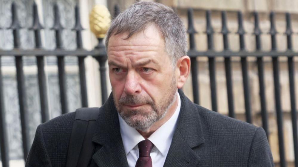 A bearded man in a white shirt, red tie and grey chequered coat frowns as he walks in front of a town hall building.