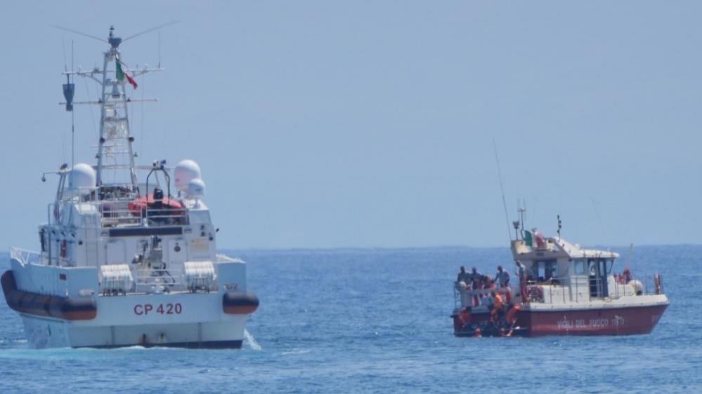 Two boats, one mainly red and the other mainly white, in the sea. with crew members visible at the stern of the smaller (red) boat.