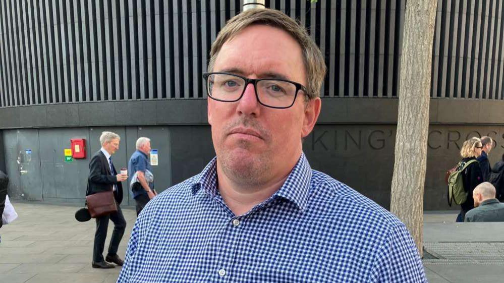 Matt Picton, a white man wearing thick-rimmed black glasses and a blue checked shirt, stands outside London's Kings Cross station