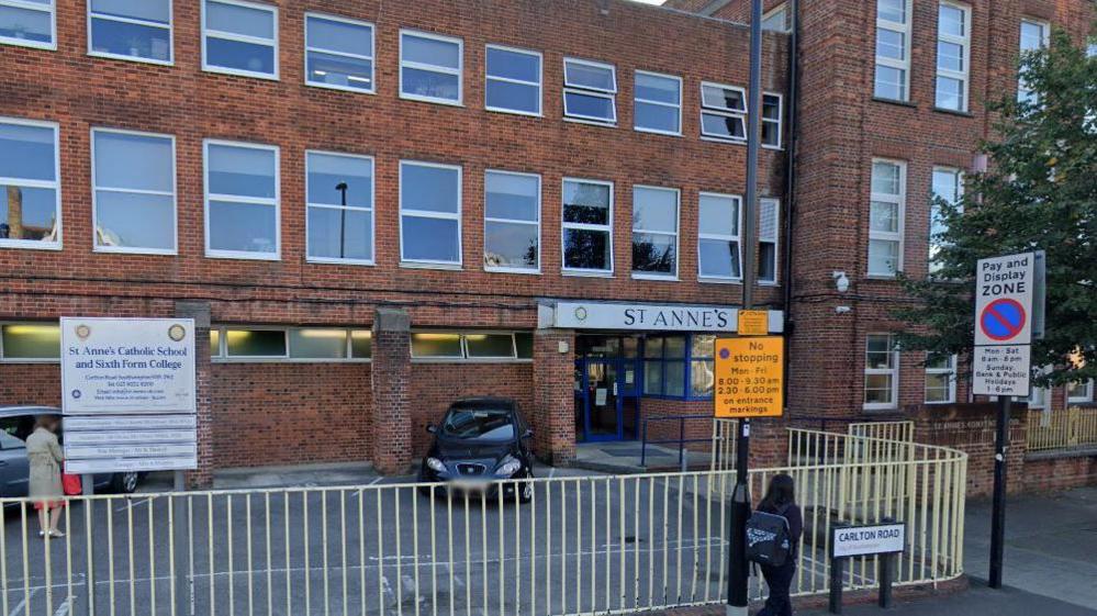 A red brick school building, behind a white rail fence. A white sign with blue writing reads St. Anne’s Catholic School and Sixth Form College.