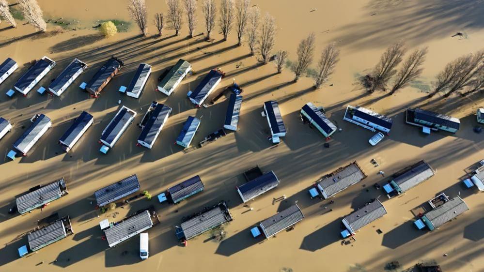 An aerial photo of a caravan park that has been flooded. Behind the caravans is a row of trees.