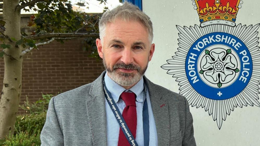 A police representative stands in front of a North Yorkshire Police sign. He is wearing a grey jacket and blue shirt, with a red tie. He has a blue lanyard around his neck. He has a short grey and black beard and grey hair. 