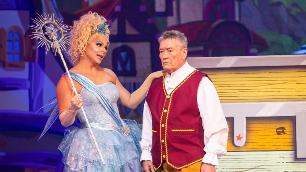 A drag queen in a sparkly blue dress stands with her hand on the shoulder of a man wearing a red vest. 