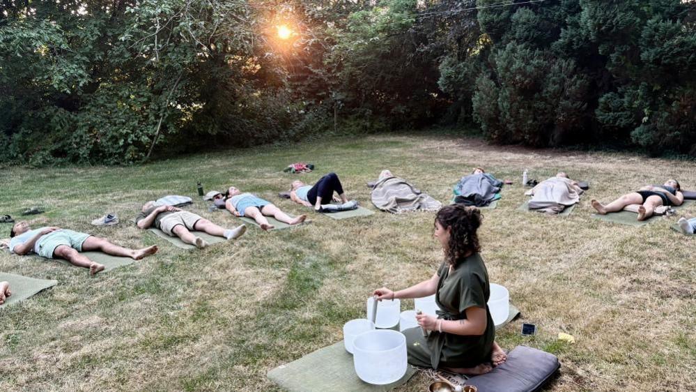A group of people are lying on the grass in a semi-circle. At the centre, a woman with long curly brown hair and wearing an olive tunic, kneels on a cushion in front of several white bowls of vafrying sizes.