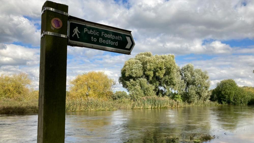 High water levels on the River Great Ouse at Kempton Mill, Bedfordshire, showing a sign that says Public Footpath to Bedford and trees and bushes along the riverbank.