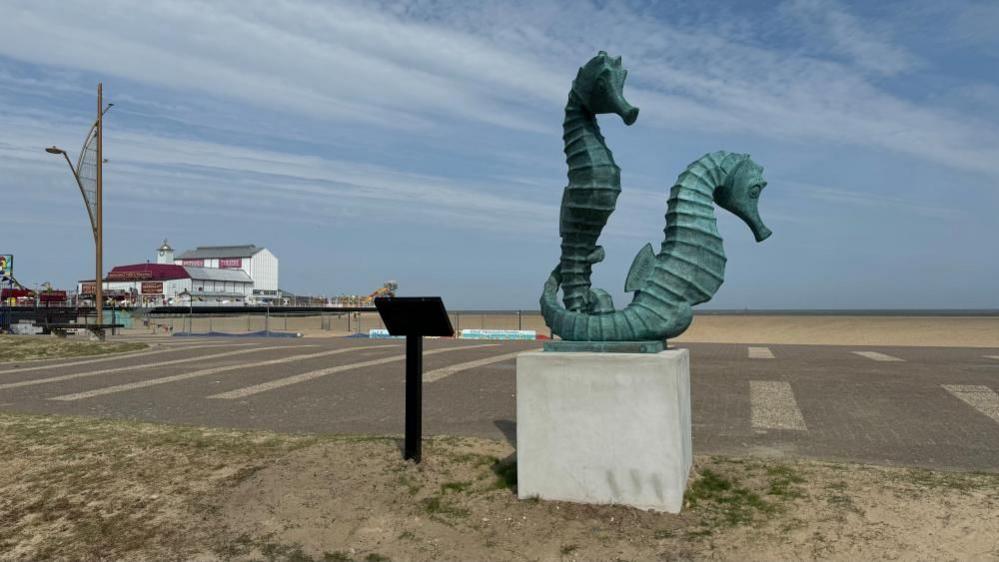 A copper-coloured sculpture of two seahorses. Behind them, the sky and sand of Great Yarmouth's Central Beach. To the left is the Britannia Pier Theatre.