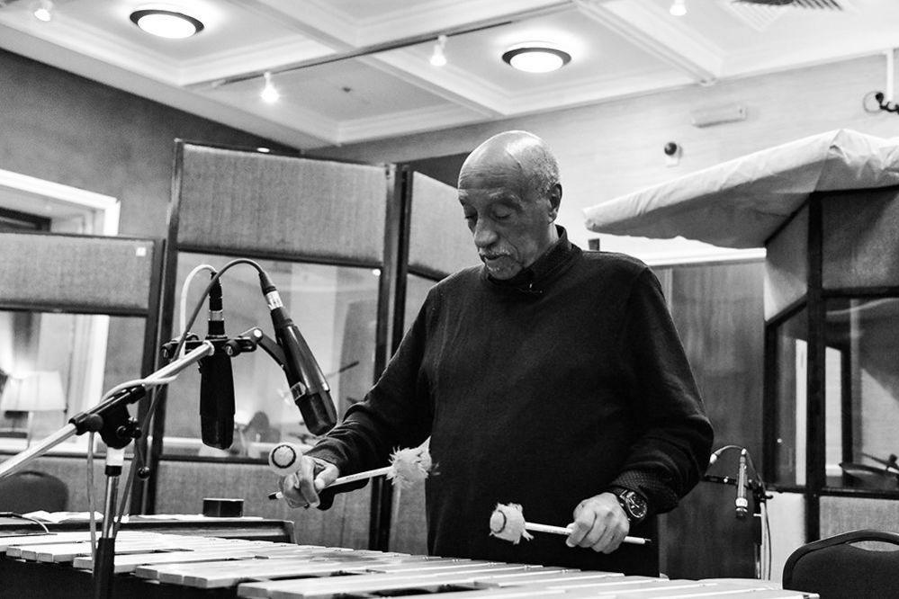 A black and white image of Mulatu Astatke in a recording studio standing at the vibraphone - he is holding the mallets as he plays the instrument.