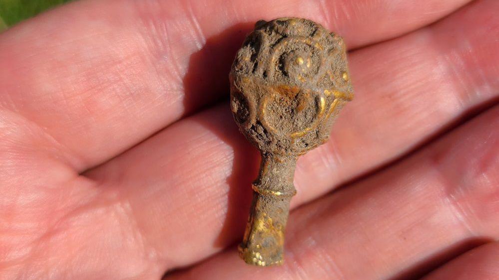 The small, gold object in the palm of someone's hand. It is covered in dirt but the bright gold can be seen underneath it. It has a stem and round top with circular patterns carved into it.