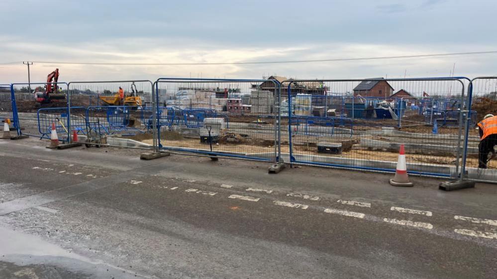 Fencing surrounds a works site where a central reservation and pedestrian crossing is being built. In the background is machinery and houses under construction.