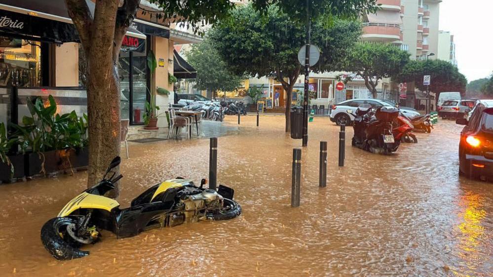 A street flooded with muddy water in Ibiza town features a yellow motorbike fallen on its side and some cars driving through the brown water.