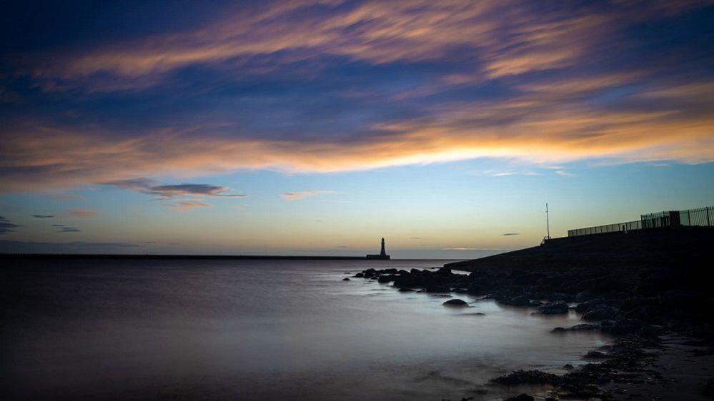 The sunrise over Roker pier which is a shadow in the distance. Wispy clouds are in the sky above and the sky is blue and orange.