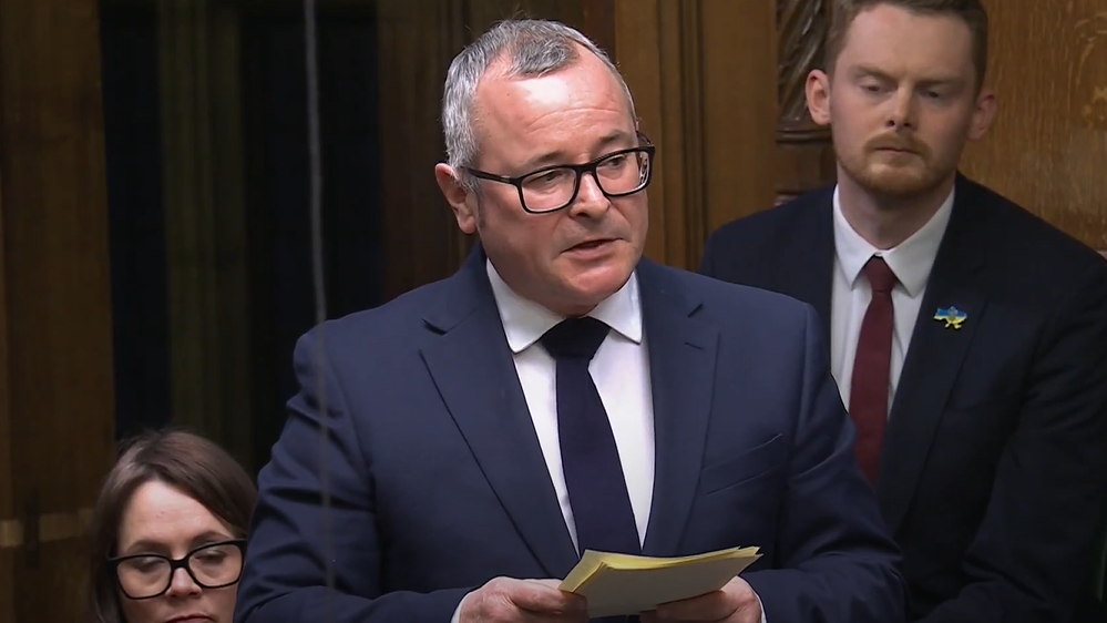 Lee Barron in a navy suit, navy tie and white shirt speaks in the House of Commons, holding some notes. 