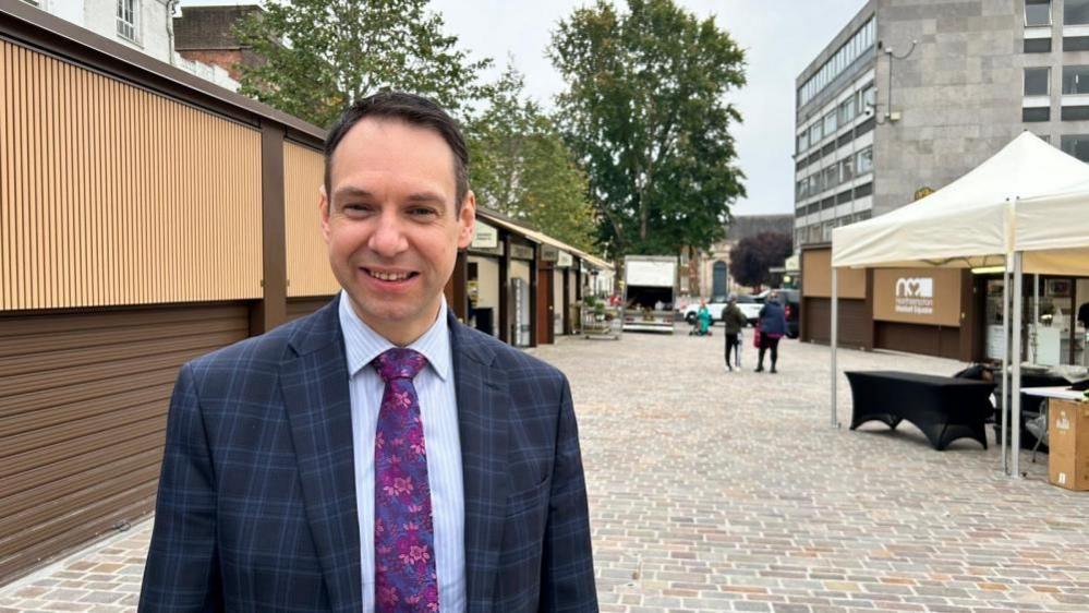 Dan Lister with short dark hair wearing a blue jacket and pink tie, standing on brick paving with market stalls behind him