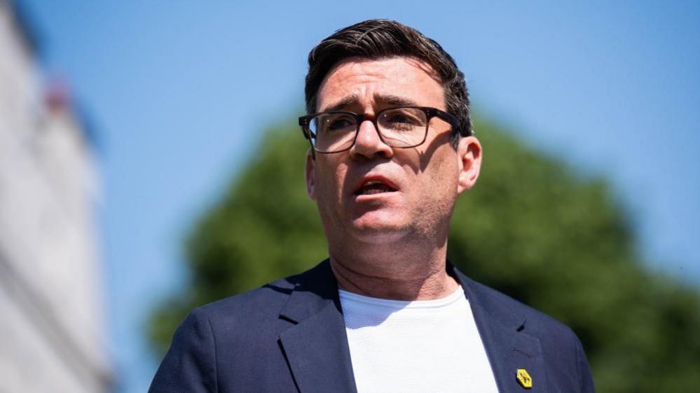 Greater Manchester Mayor Andy Burnham stands looking away from the camera. He wears glasses, a white t-shirt and a blue suit jacket. 