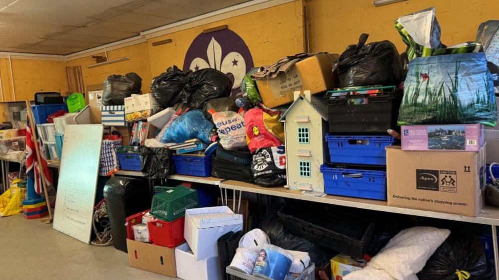A myriad of jumble in bags and boxes is stacked on and underneath tables. The wall behind is painted yellow and features a painted fleur de lys symbol of the Scouts movement.