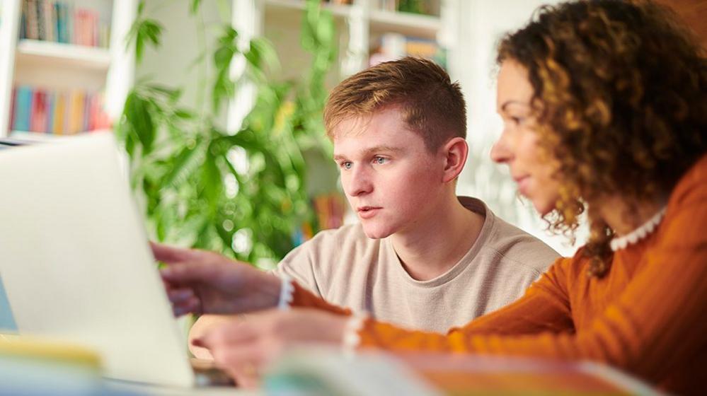 A teenage student looks at a laptop at home while being tutored by a parent or specialist