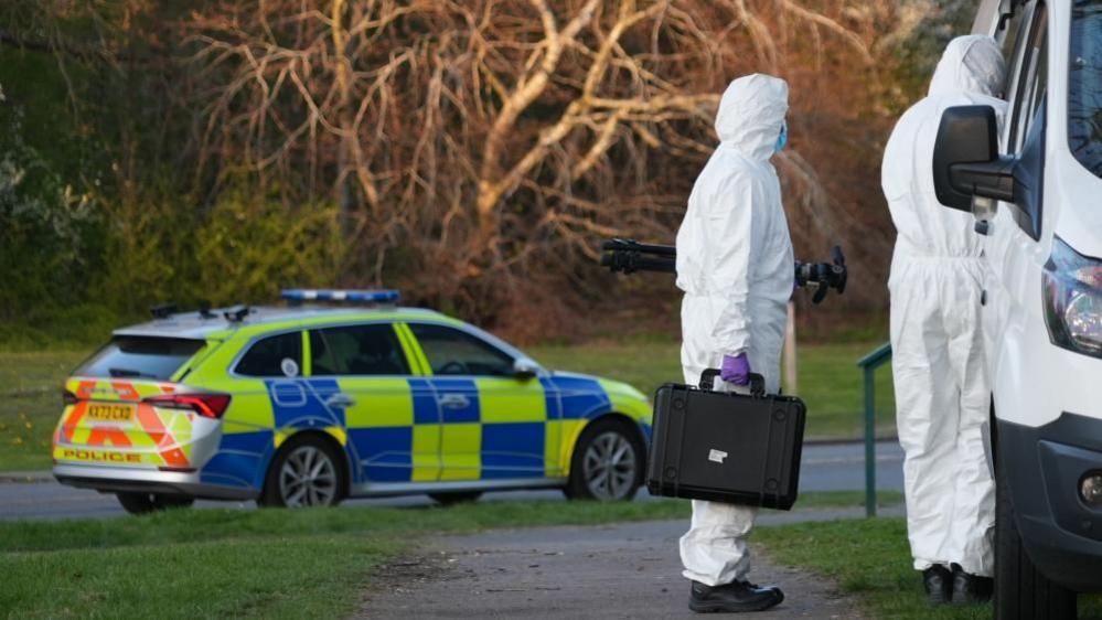 Two people in white hazmat outfits, one with a black briefcase, are standing next to a white van. There is a police car on the road to the left of the picture, with grass either side of the carriageway. There are trees in the background.