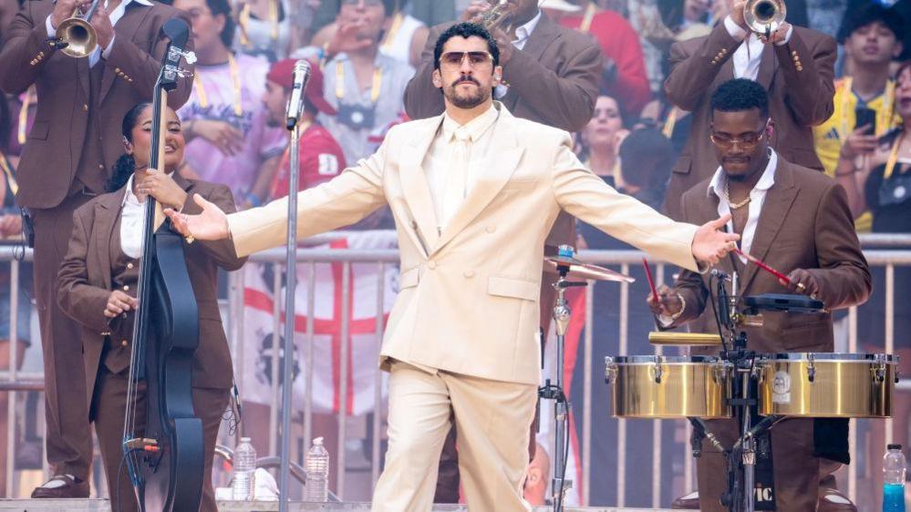 Bad Bunny in the centre with musicians surrounding him and a crowd in the background