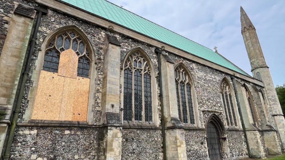 An array of the large north-facing windows on St Nicholas Minster; a trio of gothic arched window apertures, with diamond leaded glass set within, featuring a rectangular border. The windows are made of sandstone and set within a flint wall.