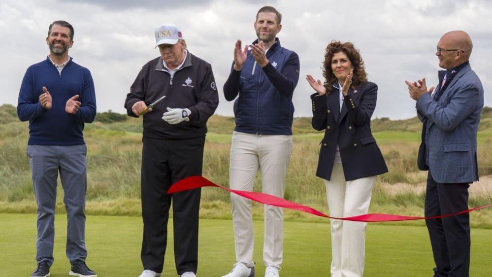 Donald Trump wearing a black golfing outfit and a white cap with a pair of scissors in his hand cutting a red ribbon which is falling to the ground. To his right is his son Donald Jr who is mid clap, wearing navy, and to this left his other son Eric wearing light coloured trousers. They are joined by a man and a woman. All have their hands clasped clapping.