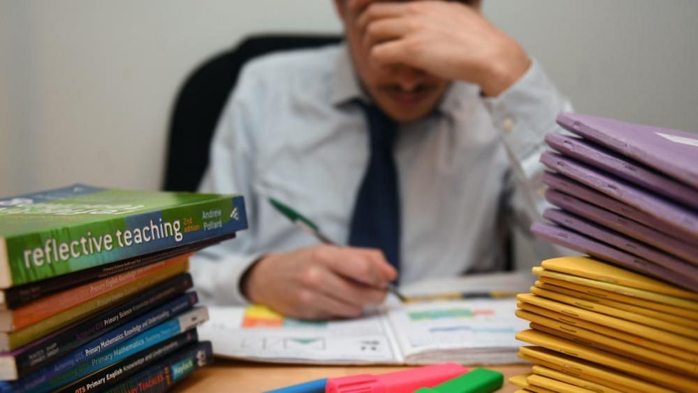 Man with short brown hair and moustache has one hand on his head and the other holding a pen with which he is writing in an exercise book.  A pile of purple and yellow books sits to his left and a pile of text books to his right.