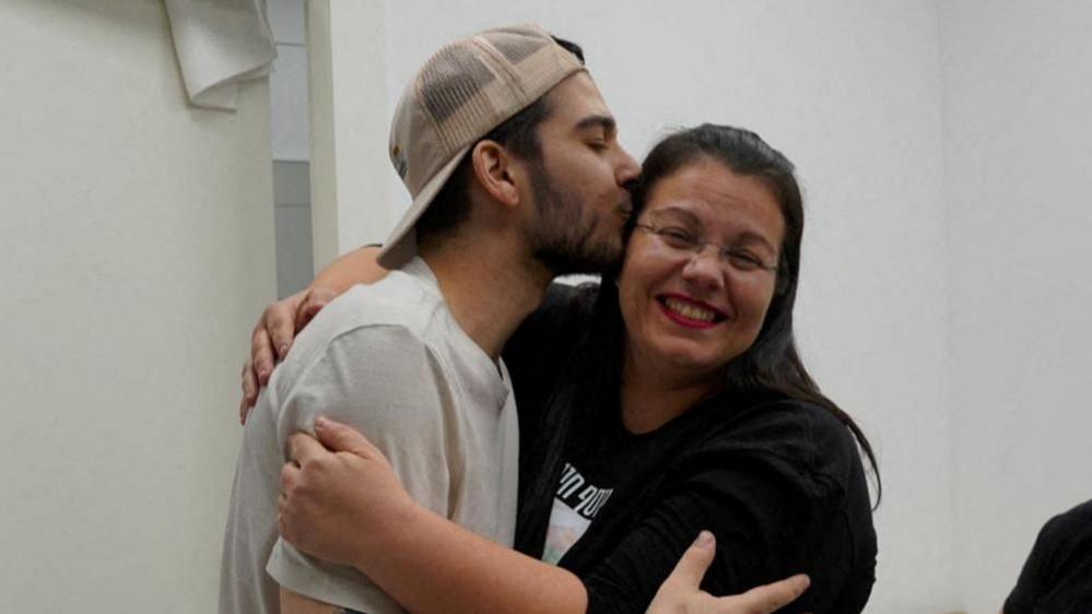 Yosef-Chaim Ohana wearing a white T-shirt and a beige baseball cap, embraces and kisses his mother who is smiling