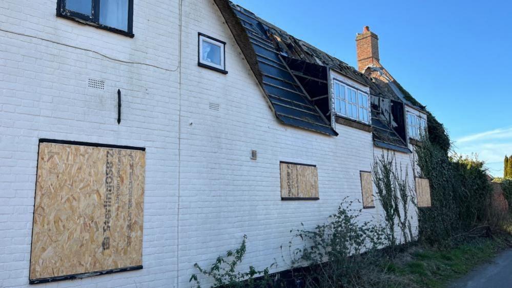 Image shows the damaged roof and boarded up windows of the house, with some vegetation in the verge near the house.