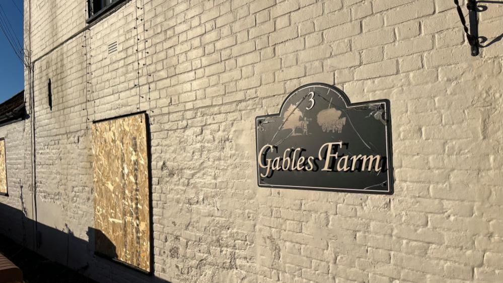 Gables Farm, pictured on a sign, on painted brickwork. The window apertures are boarded up.