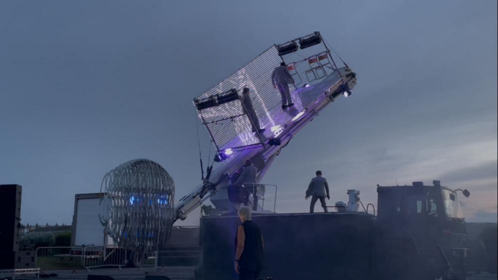 On the arm of that crane is a stage, pictured here at a 40% angle with two actors on that stage, attached by safety wire. A third cast member is on the deck of the lorry. The backdrop is the late evening sky. Ultraviolet lights illuminate the stage, which has a background netting of LED lights, with chairs mounted to the upper side panel.