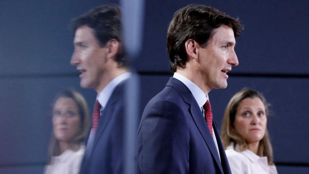 Justin Trudeau in a blue suit stands in the foreground, with Chrystia Freeland in a white shirt, looks towards him. Both are reflected in a mirrored surface behind them